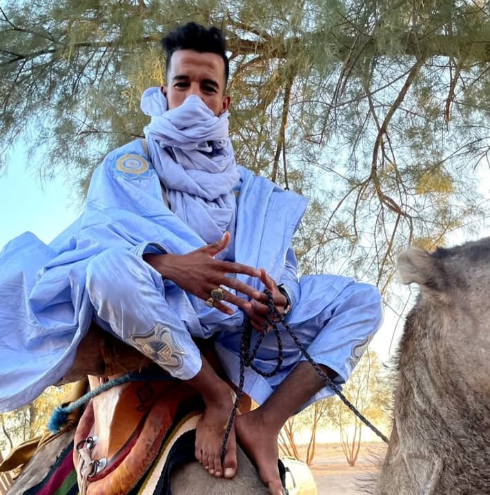 Bechir wearing traditional gandura sitting on a camel under a tamarisk tree, with a shemagh covering his nose.