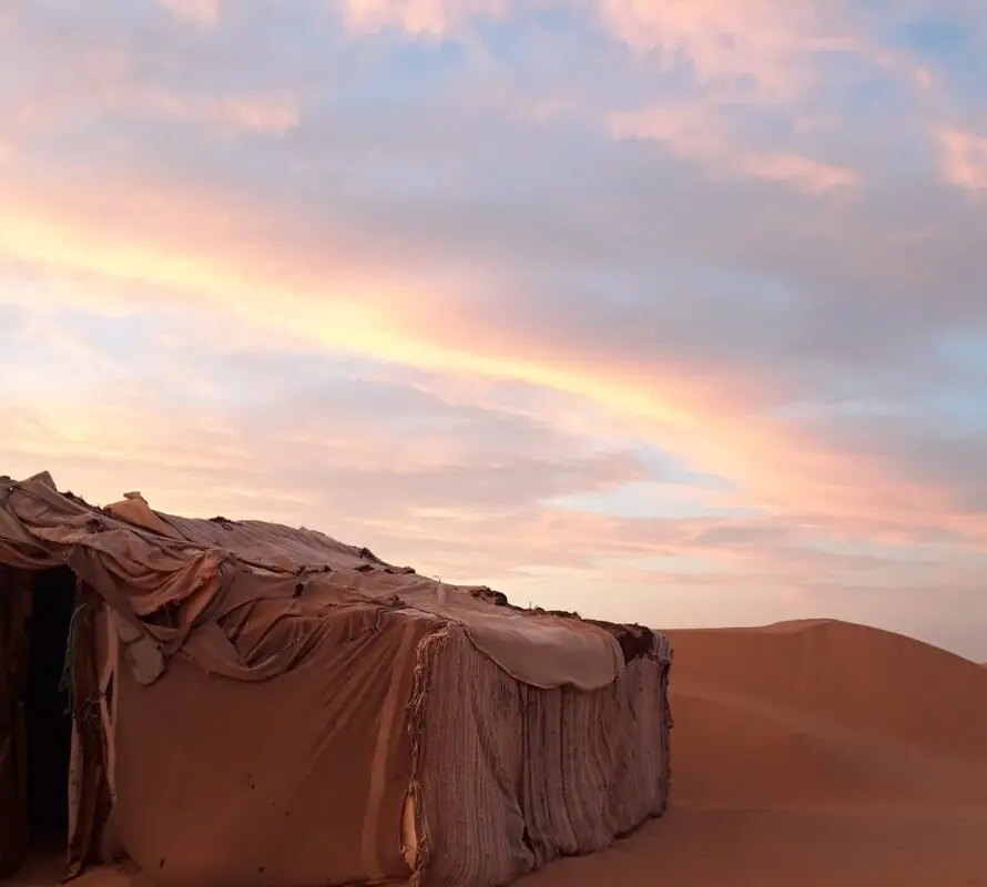 A Berber house beyond the dune under a glowing orange and grey sky — part of our nomadic Sahara desert journeys.