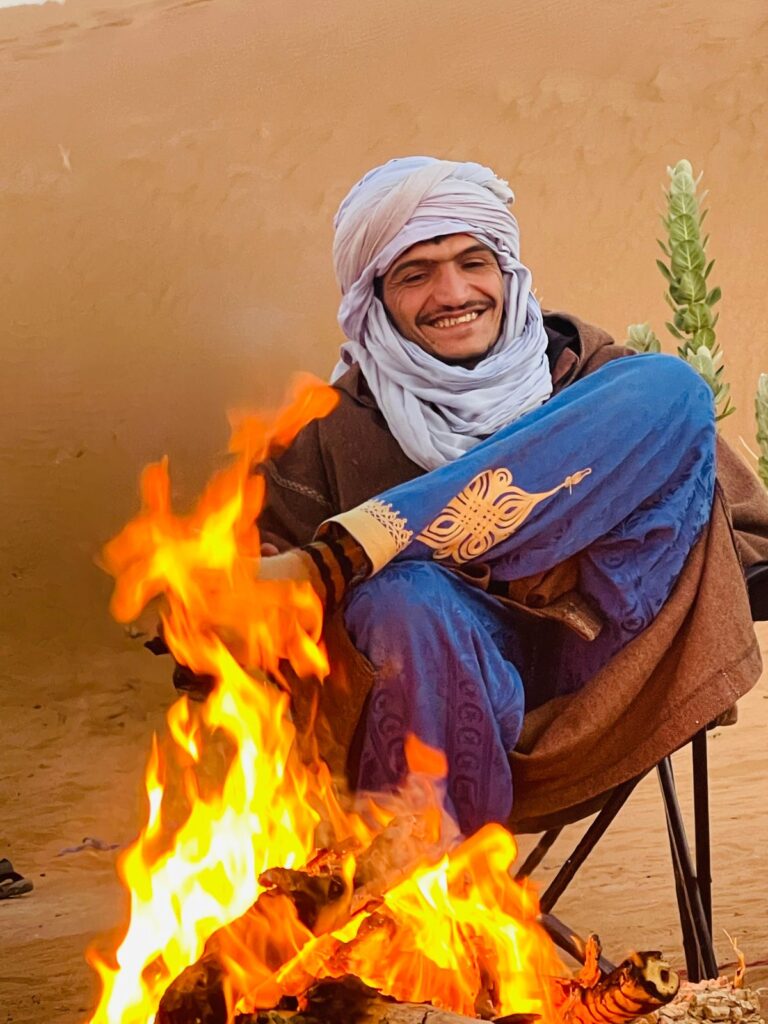 Lahsan, nomadic desert guide, sitting by a campfire in the Sahara near M'hamid, smiling in traditional Moroccan dress