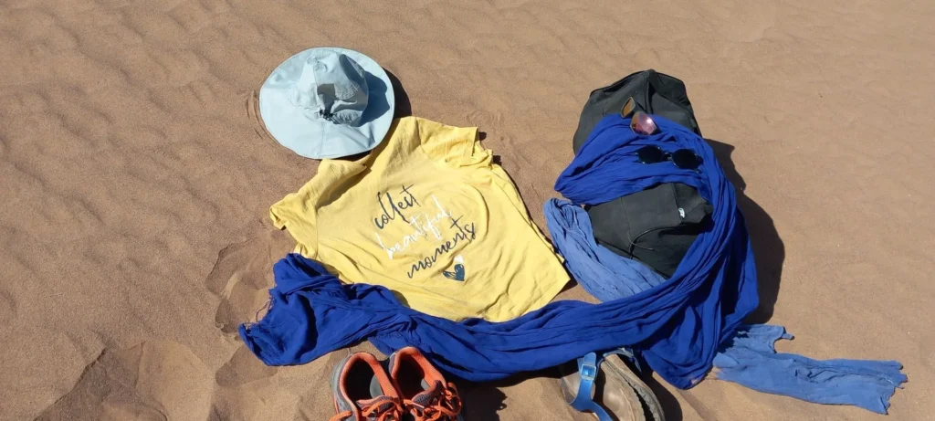 Sahara Desert packing essentials: hat, scarves, sneakers, sandals, sunglasses, and travel bag on desert sand.