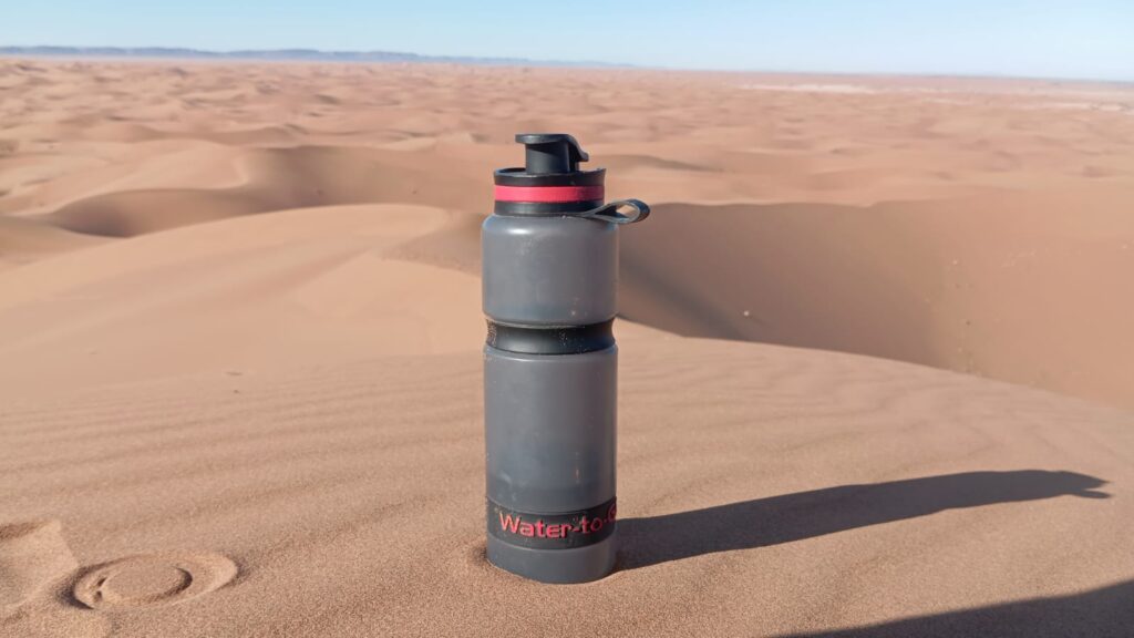 Water bottle left on the sand at the top of a dune during a trek in the Moroccan desert, with its footprint and panoramic views of the golden dunes.