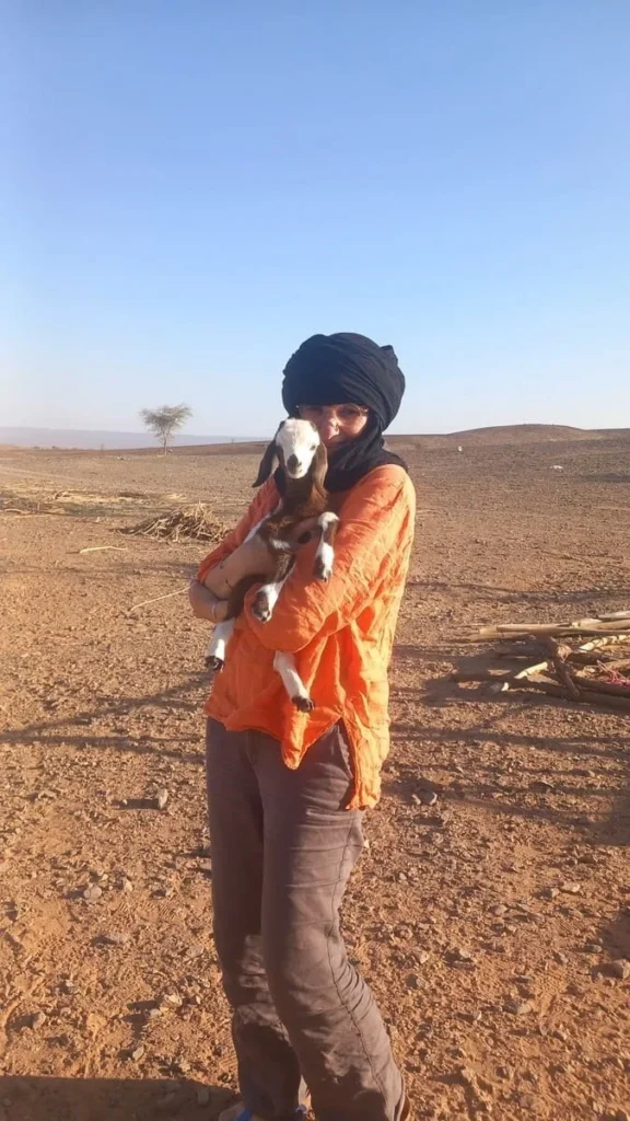 holding a kid goat against her heart, black chèche, orange shirt, chocolate linen trousers, smiling with tenderness. Acacia trees and distant mountains in the background. Arid stony ground, dry branches. Morning light, pale blue sky. Sahara Morocco.