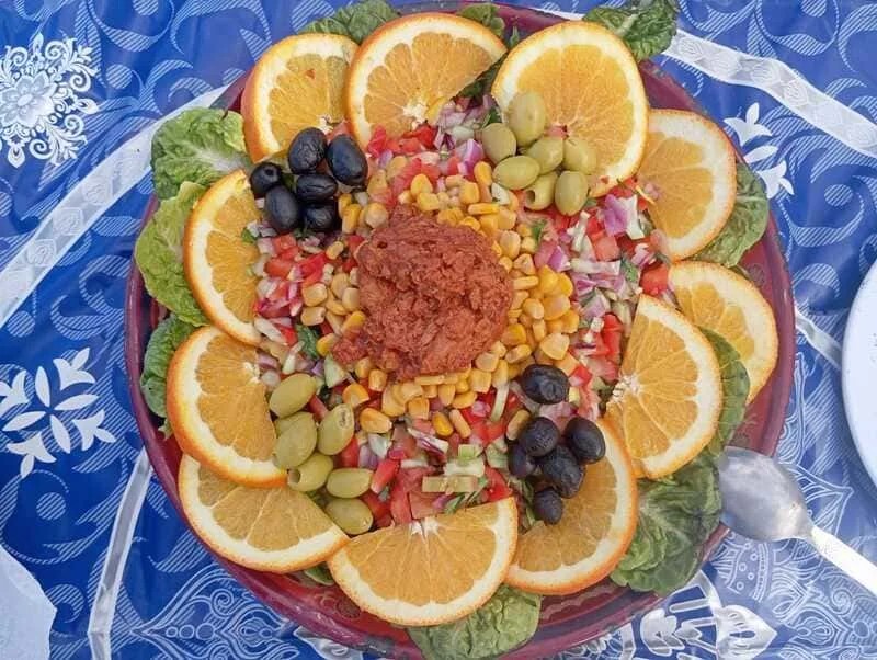 Photo of a colorful salad with fresh, crunchy vegetables served during the 6-day trek to Erg Chigaga in the Moroccan desert. Made with lettuce, orange slices, tomatoes, cucumber, coriander, corn, and green and black olives, this dish showcases the nourishing meals provided on the trek.