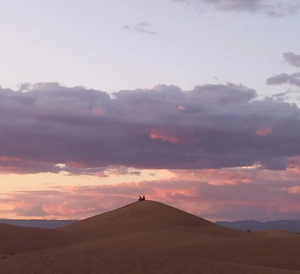 couple of trekkers silhouetted on a small dune at sunset, dramatic pink orange and pale blue cloudy sky, Shara Morocco