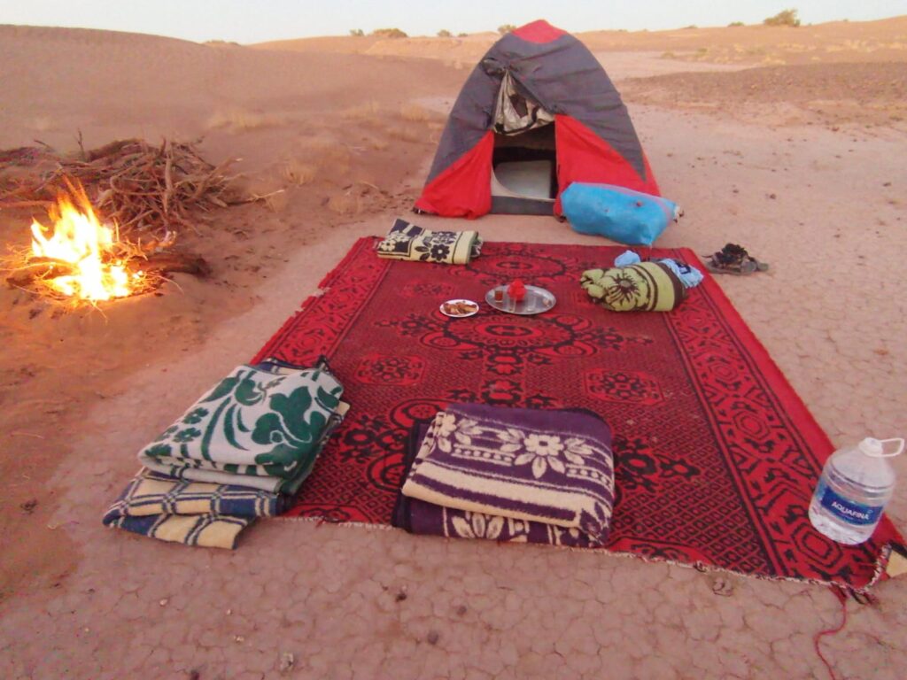 Tapis rouge traditionnel avec couvertures et thé sur un plateau, camp de trek au désert marocain, feu de bois éteint, sable et dunes sous le ciel qui s’assombrit.