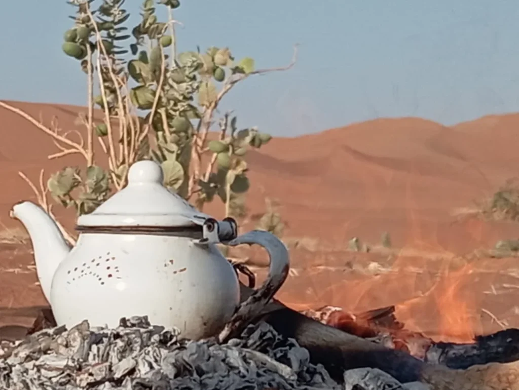 A traditional tea kettle warming over open fire in the Sahara desert, with dunes stretching into the distance near M’Hamid, Morocco.