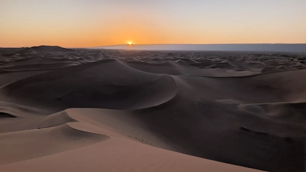 Sunset from the top of Erg Chigaga dunes, with soft shadows across the sand waves and pale pink, salmon and blue tones over the Moroccan Sahara
