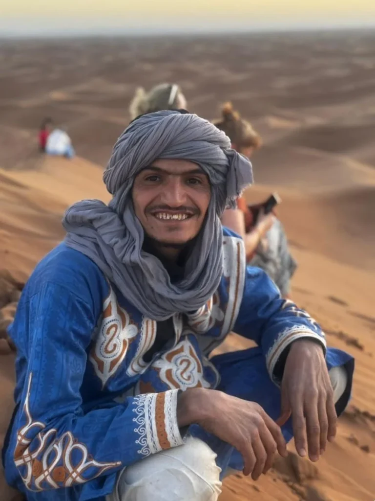 Lahsan, organisateur de retraites au Sahara, accroupi au sommet d'une dune, accueille les participants avec le sourire dans le désert marocain.